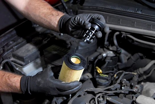 Mécanicien automobile effectuant une maintenance moteur, mains gantées manipulant un filtre à huile cylindrique neuf près du compartiment moteur ouvert
