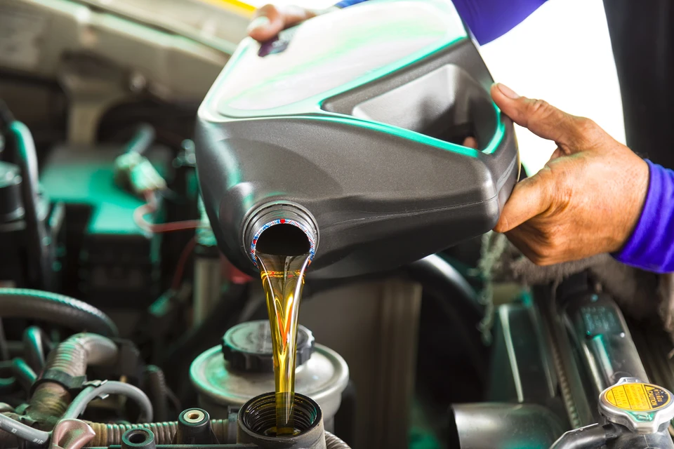 Gros plan d'une main tenant une bouteille d'additif moteur professionnel devant un moteur automobile ouvert, montrant clairement l'étiquette du produit et son versement dans le réservoir carburant ou le carter huile. L'arrière-plan doit montrer un atelier automobile professionnel. L'image doit transmettre la simplicité et la précision de l'application d'un additif pour résoudre un problème moteur spécifique.